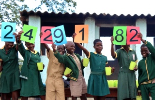 I bambini tengono in alto dei cartelli per celebrare la nuova cifra di alimentazione di Mary's Meals.